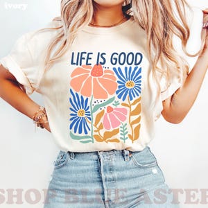 K&ouml;nnte beinhalten: Elfenbeinfarbenes T-Shirt mit dem Text "LIFE IS GOOD" &uuml;ber einem Blumendesign. Das Design zeigt orange, rosa und blaue Blumen mit gr&uuml;nen Bl&auml;ttern und Stielen. Das Shirt wird mit blauen Jeansshorts getragen.