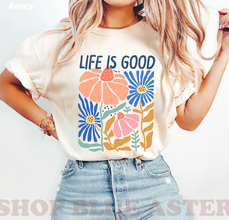 K&ouml;nnte beinhalten: Elfenbeinfarbenes T-Shirt mit dem Text "LIFE IS GOOD" &uuml;ber einem Blumendesign. Das Design zeigt orange, rosa und blaue Blumen mit gr&uuml;nen Bl&auml;ttern und Stielen. Das Shirt wird mit blauen Jeansshorts getragen.