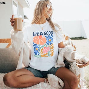 K&ouml;nnte beinhalten: Wei&szlig;es T-Shirt mit dem Text "LIFE IS GOOD" und einem bunten Blumendesign. Das Design zeigt orange, rosa und blaue Blumen mit gr&uuml;nen Bl&auml;ttern. Das Shirt ist ein l&auml;ssiges Kleidungsst&uuml;ck f&uuml;r den Alltag.