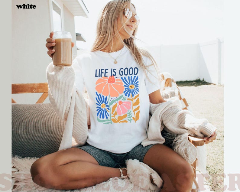 K&ouml;nnte beinhalten: Wei&szlig;es T-Shirt mit dem Text "LIFE IS GOOD" und einem bunten Blumendesign. Das Design zeigt orange, rosa und blaue Blumen mit gr&uuml;nen Bl&auml;ttern. Das Shirt ist ein l&auml;ssiges Kleidungsst&uuml;ck f&uuml;r den Alltag.