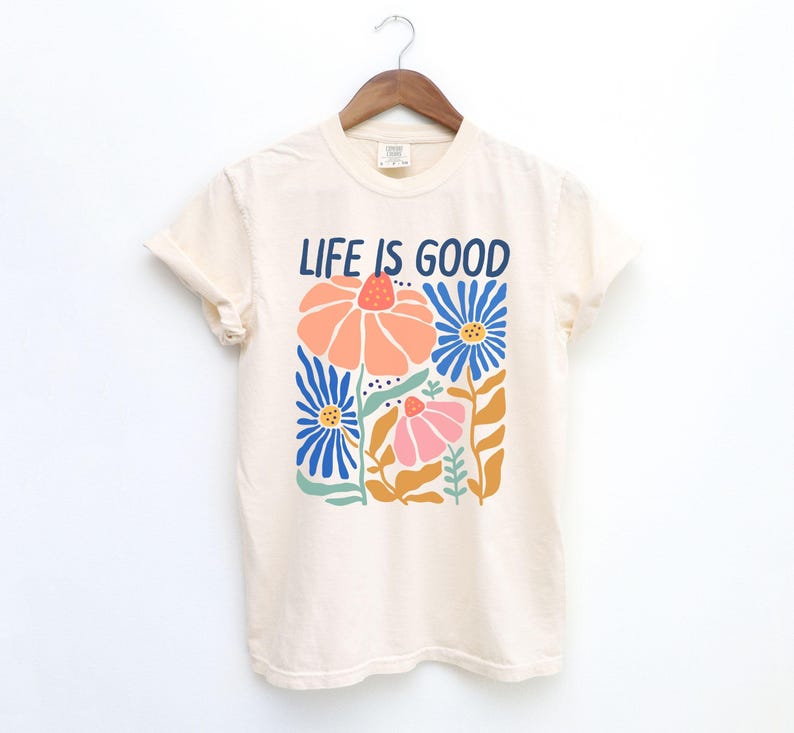 K&ouml;nnte beinhalten: Cremefarbenes T-Shirt mit dem Schriftzug "LIFE IS GOOD" in Marineblau. Das Shirt zeigt ein Blumendesign mit orangefarbenen, blauen und rosa Blumen. Das T-Shirt h&auml;ngt an einem Holzb&uuml;gel.