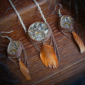 Puede incluir: Juego de joyas estilo steampunk con collar y pendientes. El collar tiene un colgante redondo con engranajes de reloj visibles, una pluma de cobre y un borde con cuentas. Los pendientes reflejan el diseño del colgante, cada uno con una pluma de cobre.