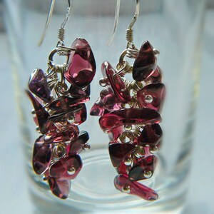 May include: A pair of dangle earrings featuring clusters of deep red-purple gemstones. The earrings are suspended from silver-colored hooks and connected by silver-toned wire. The gemstones are irregularly shaped and clustered together, creating a textured appearance.
