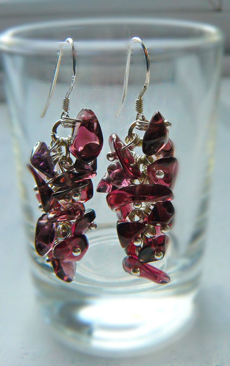May include: A pair of dangle earrings featuring clusters of deep red-purple gemstones. The earrings are suspended from silver-colored hooks and connected by silver-toned wire. The gemstones are irregularly shaped and clustered together, creating a textured appearance.