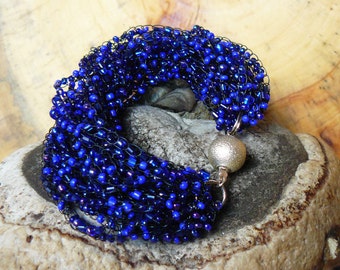 Peut inclure: Un bracelet artisanal composé de perles bleu foncé tissées ensemble. Le bracelet est doté d'un fermoir magnétique argenté. Les perles, de formes variées, créent un effet texturé. Le bracelet mesure environ 17 cm de circonférence.