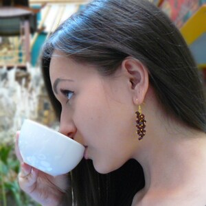 May include: A woman wearing gold-colored earrings with a cluster of dark red beads. The earrings are dangling. She is holding a white cup and drinking from it. The background is blurred.