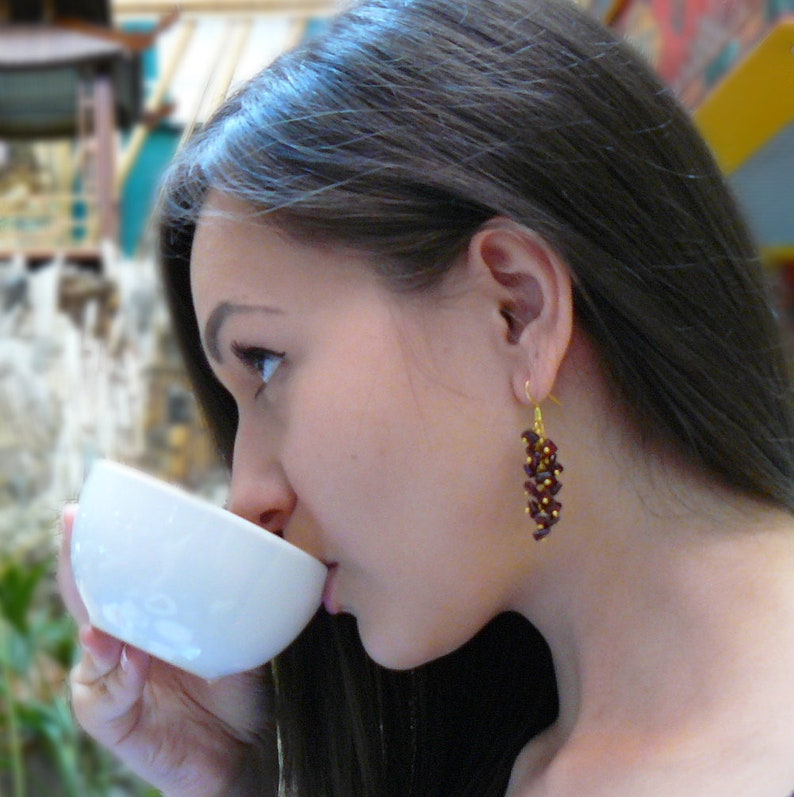 May include: A woman wearing gold-colored earrings with a cluster of dark red beads. The earrings are dangling. She is holding a white cup and drinking from it. The background is blurred.