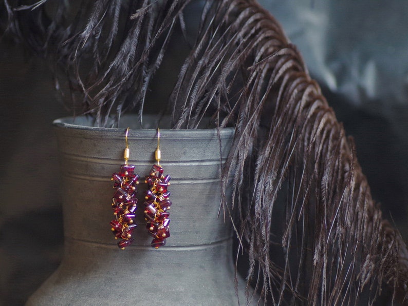 May include: A pair of dangle earrings with a cluster design. The earrings feature a deep red color with gold-colored accents. The earrings are suspended from gold-colored hooks. The background includes a dark gray metal container and dark feathers.