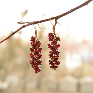 May include: A pair of dangle earrings with a cluster of deep red, faceted gemstones. The earrings are suspended from gold-colored hooks and are arranged in a cascading design. The background is softly blurred, highlighting the jewelry.