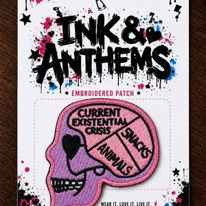 May include: A purple skull-shaped embroidered patch on a white backing card with black, pink, and blue paint splatter designs. The patch features a brain diagram labelled with the text Current Existential Crisis, Animals, and Snacks in black.