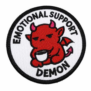 May include: A circular embroidered patch with a white background and a black border. It features a red cartoon demon holding a white cup. The text EMOTIONAL SUPPORT DEMON is embroidered in black capital letters along the top and bottom edges.
