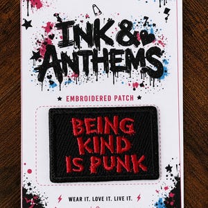 May include: A rectangular black embroidered patch with red stitched text that reads BEING KIND IS PUNK. The patch is mounted on a white display card featuring black and blue paint splatter designs, stars, and the text INK & ANTHEMS.