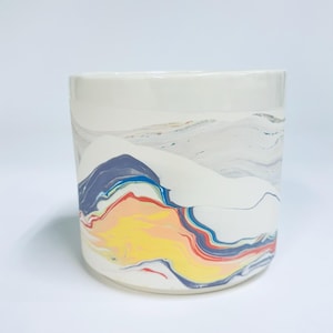 May include: A cylindrical ceramic planter featuring a white base with a marbled design. The surface displays swirling patterns of yellow, red, blue, and grey paint, creating a fluid, multi-coloured effect across the exterior of the vessel.