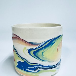 May include: A cylindrical ceramic planter featuring a marbled pattern with swirls of blue, lime green, and soft peach colours against an off-white base. The vessel has a smooth, straight-sided exterior and a wide, open top with a clean rim.