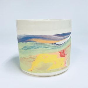 Peut inclure: Un pot de fleurs cylindrique en céramique avec un bord supérieur blanc lisse et une section inférieure marbrée aux motifs tourbillonnants de bleu, jaune, vert et rouge. Le récipient a une base plate et des parois droites multicolores.