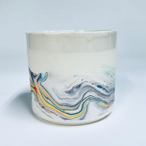 May include: A cylindrical ceramic mug featuring a smooth, light grey glazed rim and a white base decorated with a swirling, marbled pattern of blue, yellow, red, and grey paint. The vessel has a straight-sided profile and a flat bottom edge.