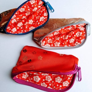 May include: Three small, open zippered pouches. The exterior fabrics are gold, bronze, and red. The interiors are lined with red fabric featuring a white floral pattern. The zippers are blue, silver, and purple.