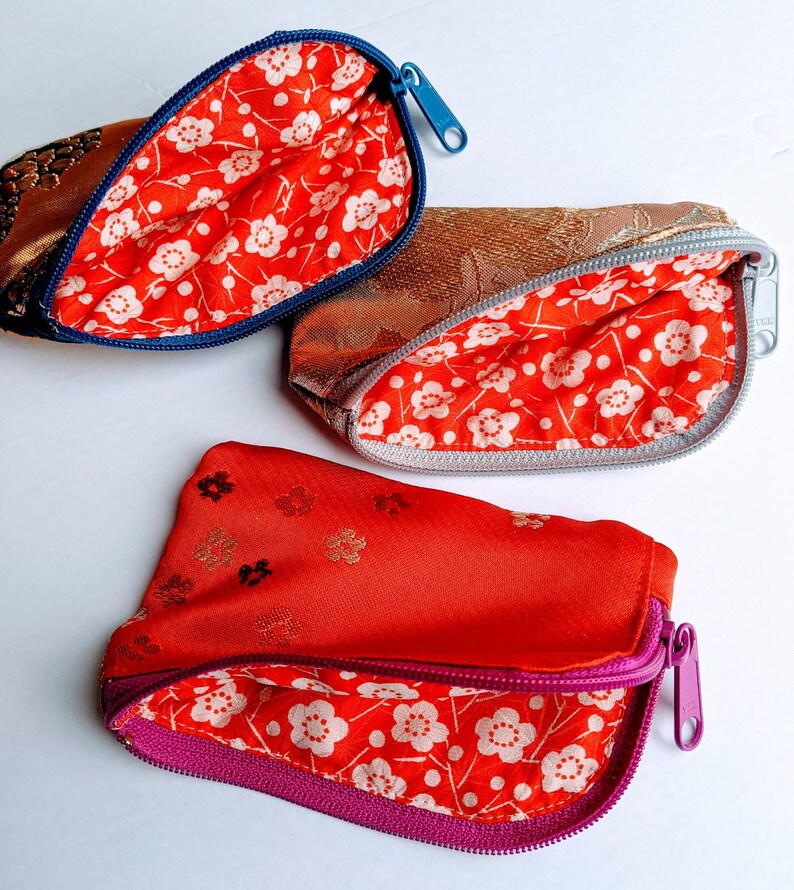 May include: Three small, open zippered pouches. The exterior fabrics are gold, bronze, and red. The interiors are lined with red fabric featuring a white floral pattern. The zippers are blue, silver, and purple.
