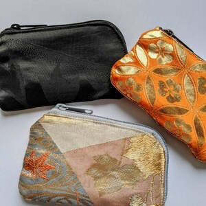 May include: Three small zippered pouches in the image. One is black, one is orange with a gold floral pattern, and the third is a patchwork of gold, pink, and blue fabrics with a leaf design. All have a rectangular shape.