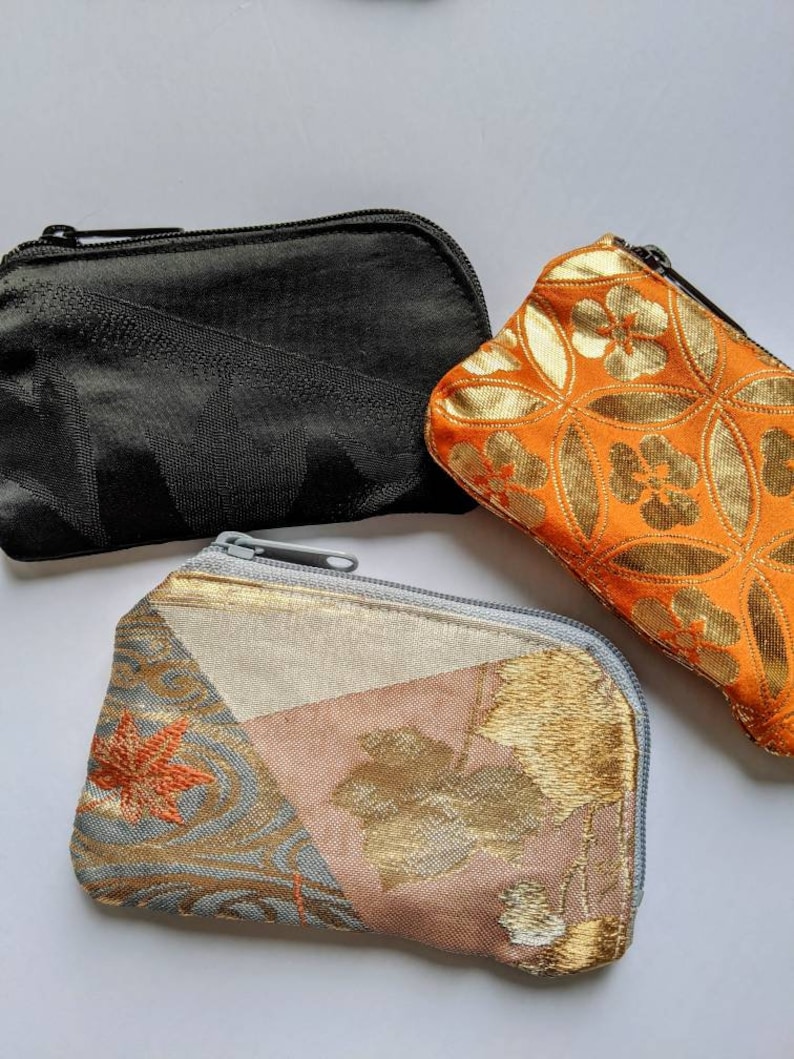 May include: Three small zippered pouches in the image. One is black, one is orange with a gold floral pattern, and the third is a patchwork of gold, pink, and blue fabrics with a leaf design. All have a rectangular shape.