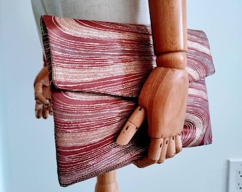 May include: A burgundy and gold clutch with a swirling pattern. The rectangular bag has a fold-over flap and is held by a wooden mannequin hand. The bag's design features horizontal lines of burgundy and gold.
