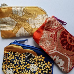 May include: Three small, zippered pouches in a variety of colors and patterns. One is gold and cream with a geometric design, another is red and orange with a floral pattern, and the third is navy blue with gold accents.