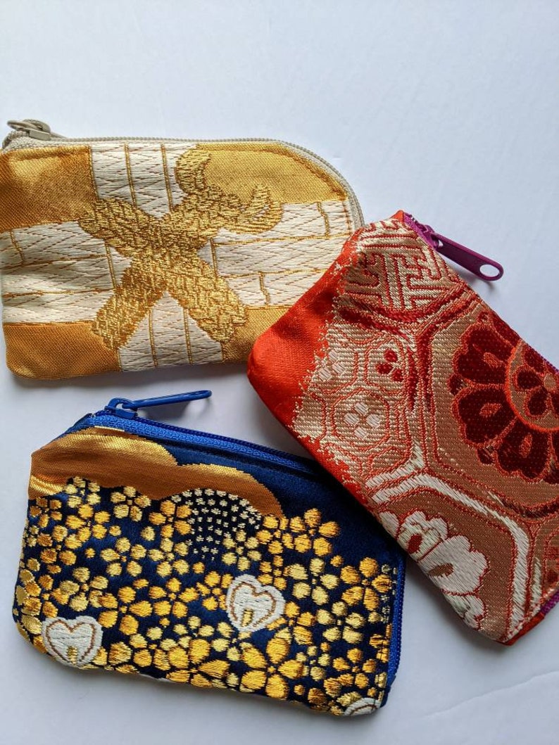 May include: Three small, zippered pouches in a variety of colors and patterns. One is gold and cream with a geometric design, another is red and orange with a floral pattern, and the third is navy blue with gold accents.