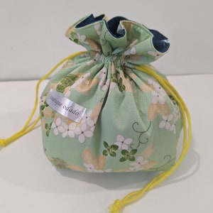 Op de afbeelding: Een kleine zak met trekkoord en een bloemenpatroon in groen, wit en geel. De zak heeft een gele trekkoord en een label met de tekst "boutique odado". De binnenvoering is donkerblauw.