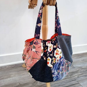 May include: A patchwork tote bag with floral and geometric patterns in pink, navy, and grey. The bag has navy blue straps and a red interior. The bag is displayed on a wooden stand.