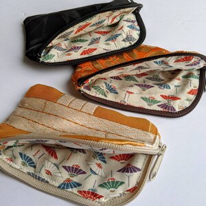 May include: Three small, open zippered pouches in black, orange, and tan. The pouches have a patterned interior with colorful parasols. The tan pouch has a quilted design. The black pouch has a diamond pattern.