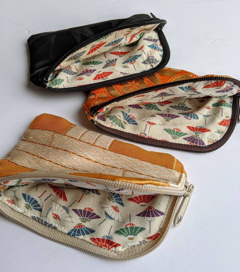 May include: Three small, open zippered pouches in black, orange, and tan. The pouches have a patterned interior with colorful parasols. The tan pouch has a quilted design. The black pouch has a diamond pattern.