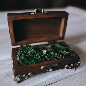 A wooden ring box with a hinged lid and metal accents. The box is open and filled with green moss.