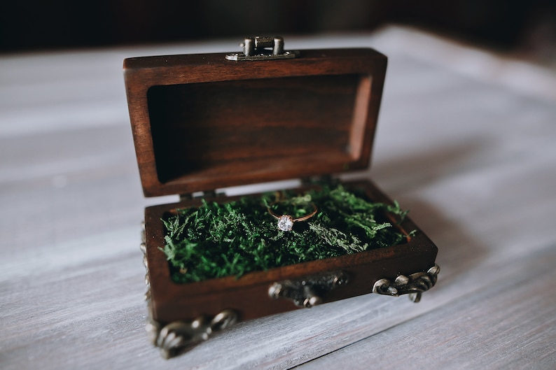 A wooden ring box with a hinged lid and metal accents. The box is open and filled with green moss.