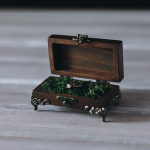 A wooden ring box with a hinged lid, open to reveal on a bed of green moss.