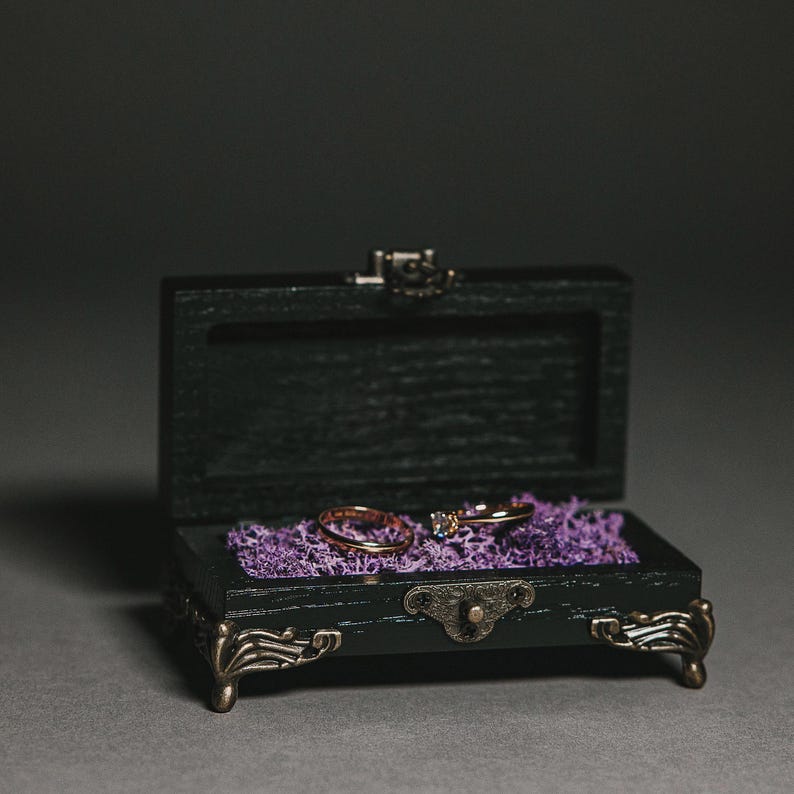 The sleek black finish gives this wooden ring box a modern yet timeless appeal, perfect for any wedding theme. The contrasting metallic hardware adds a touch of vintage charm.