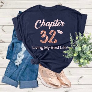 May include: Navy blue t-shirt with rose gold glitter text that reads "Chapter 32 Living My Best Life". A pink lip print is above the number 32.