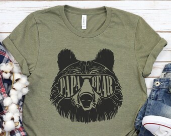 May include: A green t-shirt with a black bear wearing sunglasses and the text "Papa Bear". The t-shirt is laid out on a wooden surface with a plaid shirt, cotton balls, a pair of jeans, and white sneakers.