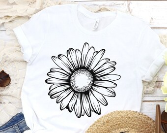 May include: A white t-shirt with a black and white daisy graphic. The shirt is laid out on a white wooden surface with a pair of brown sunglasses, a pair of brown sandals, a straw hat, and a white lace shawl.