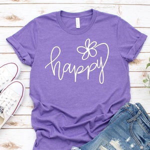 May include: Purple t-shirt with a white graphic that says "happy" with a flower design. The shirt is laying on a white wooden surface with a pair of white sneakers and denim shorts.