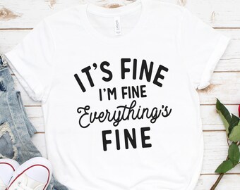 May include: White t-shirt with black text that reads "It's Fine I'm Fine Everything's Fine". The shirt is laying on a white wooden surface with a pair of white sneakers, blue jeans, and a red rose.