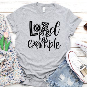 Peut inclure: Un t-shirt gris avec le texte "Lead by example" imprimé en noir. Le "A" du mot "Lead" est un motif rayé noir et blanc.
