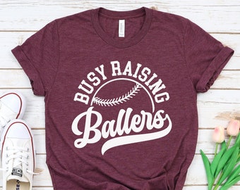 Può includere: Una maglietta color granata con il testo "Busy Raising Ballers" in bianco, con una grafica di baseball. La maglietta è su uno sfondo di legno bianco con scarpe da ginnastica bianche e fiori rosa.