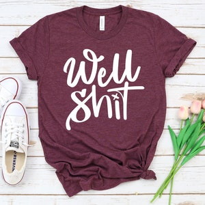 May include: A maroon t-shirt with the phrase "Well Shit" written in white cursive lettering.