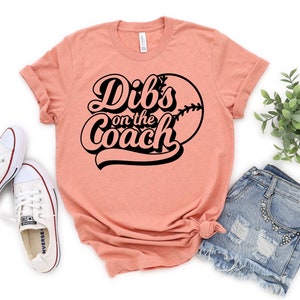 May include: A peach-colored t-shirt with black text that reads "Dibs on the Coach" and a baseball graphic.