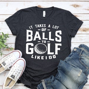 May include: A dark gray t-shirt with white text that reads "It takes a lot of balls to golf like I do". A white golf ball is pictured in the text. The shirt is laid out on a white wood surface with a pair of white sneakers and blue jeans.
