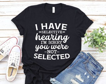 Może przedstawiać: Czarny T-shirt z białym tekstem, który brzmi "Mam selektywny słuch, przepraszam, nie zostałeś wybrany". Koszulka leży na drewnianej powierzchni z parą białych butów sportowych i niebieskimi dżinsowymi spodenkami.