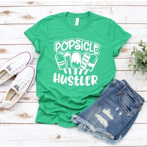 May include: Green t-shirt with white text that reads "Popsicle Hustler" and a graphic of five popsicles. The t-shirt is laying on a white wooden surface with a pair of white sneakers and denim shorts.