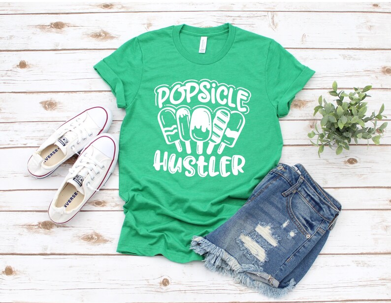 May include: Green t-shirt with white text that reads "Popsicle Hustler" and a graphic of five popsicles. The t-shirt is laying on a white wooden surface with a pair of white sneakers and denim shorts.