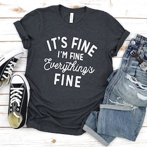 May include: A dark gray t-shirt with white text that reads "It's Fine I'm Fine Everything's Fine". The shirt is laid flat on a white wooden surface with a pair of black sneakers and a pair of blue jeans.