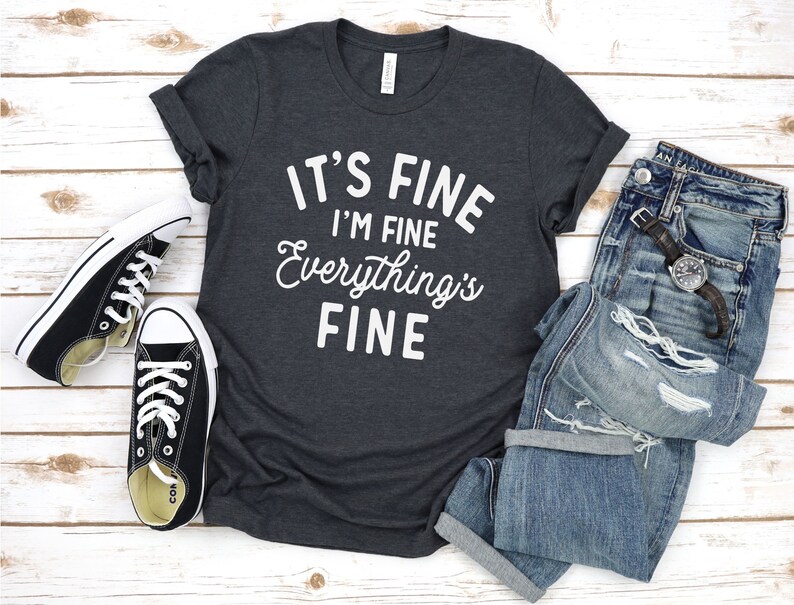 May include: A dark gray t-shirt with white text that reads "It's Fine I'm Fine Everything's Fine". The shirt is laid flat on a white wooden surface with a pair of black sneakers and a pair of blue jeans.